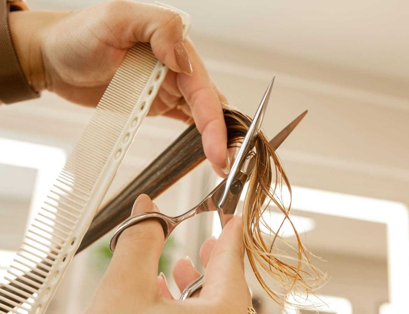 Hands using scissors and comb to cut brown hair in a salon setting.