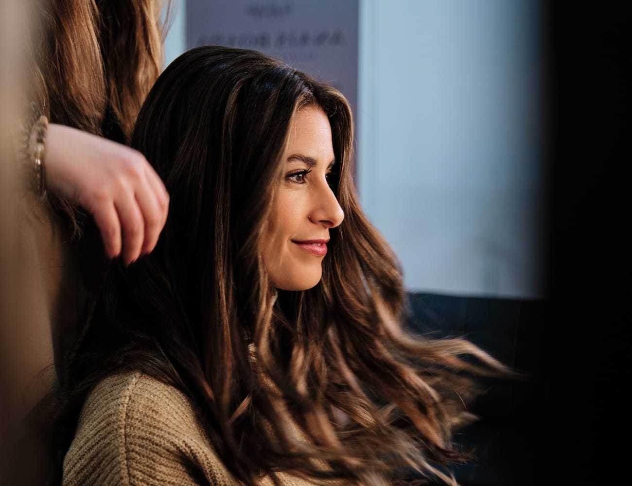 Woman with long hair in a salon, smiling as her hair is styled.
